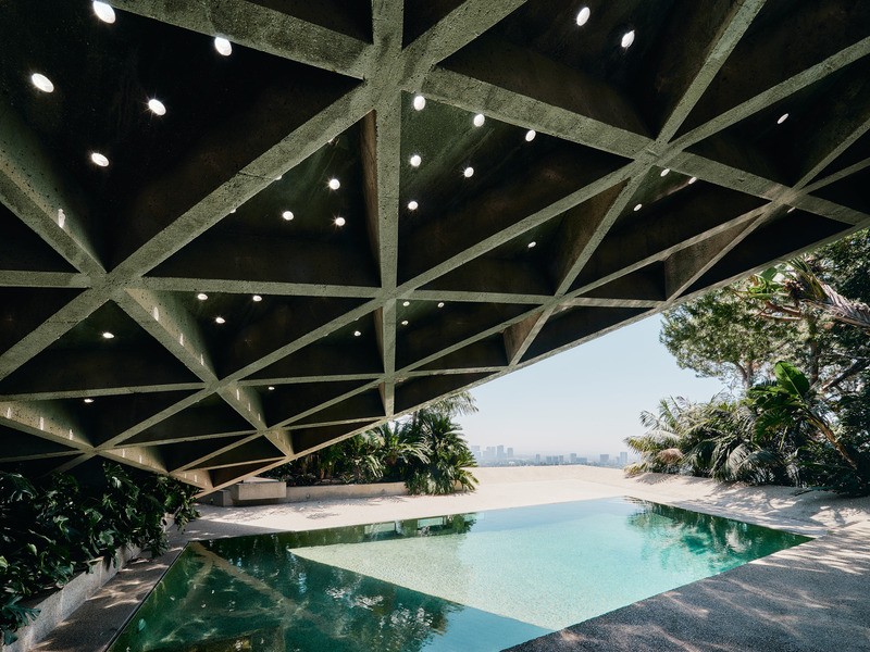 Vue de la salle de séjour sur la terrasse de la piscine enveloppée de béton fini par des galets. Le toit à caissons en béton triangulé, à la fois puissant et léger, est suspendu au-dessus de la tête, ponctué de minuscules lucarnes qui laissent passer la l